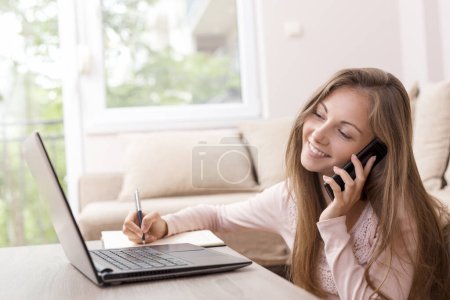 Photo for Young female student studying and exchanging notes with her colleagues using modern technologies, having a phone conversation and taking notes - Royalty Free Image