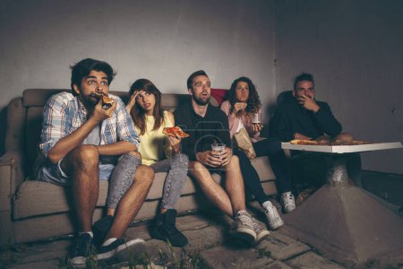 Téléchargez les photos : Groupe de jeunes amis regardant un film d'horreur sur un toit-terrasse du bâtiment, mangeant de la pizza, buvant de la bière et s'amusant - en image libre de droit