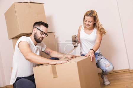 Foto de Chica joven mudándose en un nuevo apartamento con su novio, de pie rodeado de cajas de cartón, embalaje y cajas de cinta, mientras que el novio lleva cajas de distancia - Imagen libre de derechos