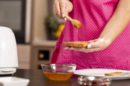 Foto de Detalle de manos femeninas sosteniendo rebanada de pan tostado y cuchara de madera, extendiendo miel sobre el pan; mujer haciendo mantequilla y sándwich de miel para el desayuno - Imagen libre de derechos