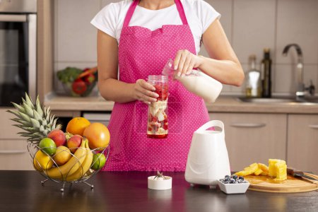 Téléchargez les photos : Détail d'une femme versant du lait sur les fruits dans un bol mélangeur et faisant framboise fraîche, banane, lait et smoothie au miel pour le petit déjeuner - en image libre de droit