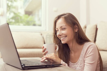 Téléchargez les photos : Jeune belle femme assise dans le salon, buvant du café et utilisant un ordinateur portable - en image libre de droit