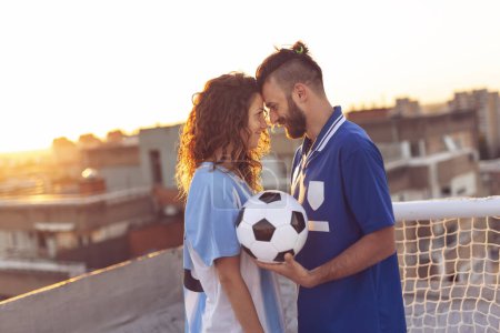Téléchargez les photos : Couple amoureux portant des maillots de football, debout sur le toit d'un immeuble après un match et profitant d'un magnifique coucher de soleil sur la ville - en image libre de droit