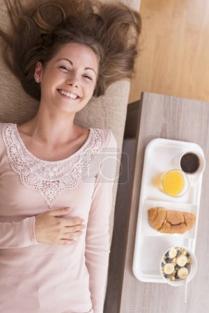 Foto de Vista superior de una hermosa joven tumbada en un sofá con bandeja que contiene deliciosa comida para el desayuno colocada junto a ella - Imagen libre de derechos