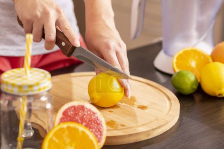 Photo for Detail of female hands holding a kitchen knife and cutting lemon on a cutting board; woman making a freshly squeezed mixed citrus fruit on a kitchen counter - Royalty Free Image