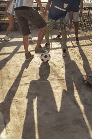Foto de Grupo de jóvenes amigos con camisetas jugando al fútbol en una terraza en la azotea de un edificio en un día soleado de verano, detalle de piernas pateando la pelota - Imagen libre de derechos