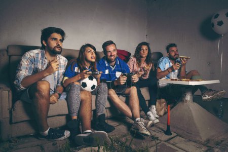 Foto de Grupo de jóvenes amigos divirtiéndose en una terraza en la azotea mientras juegan un videojuego de fútbol - Imagen libre de derechos