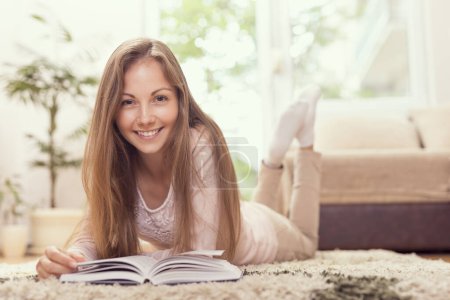 Téléchargez les photos : Jolie jeune femme couchée sur le sol à la maison et lisant un livre - en image libre de droit