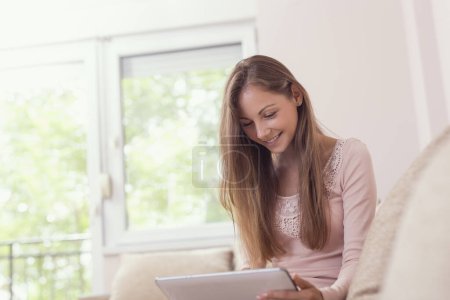 Téléchargez les photos : Belle jeune fille assise sur un canapé du salon, tenant une tablette et surfant sur le net - en image libre de droit