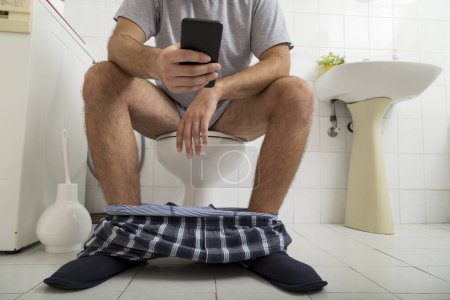 Téléchargez les photos : Détail d'un homme assis sur les toilettes avec son pyjama et ses sous-vêtements, lisant le journal sur un téléphone intelligent - en image libre de droit