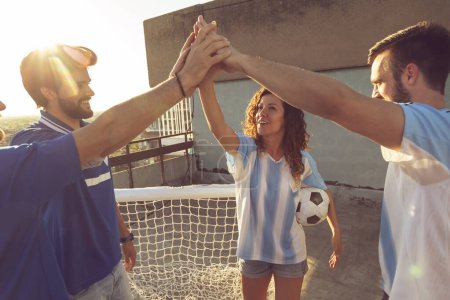 Foto de Grupo de jóvenes amigos divirtiéndose jugando al fútbol en una terraza de la azotea del edificio, saludando antes del comienzo del partido, haciendo grupo de cinco - Imagen libre de derechos
