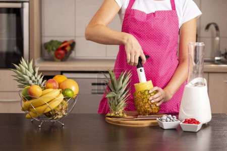 Téléchargez les photos : Femme épluchant un ananas avec un coupe-ananas, faisant un smoothie de fruits frais, sains et crus - en image libre de droit