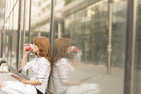 Téléchargez les photos : Jeune femme d'affaires pendant une pause café à l'extérieur de son bureau boire du café et travailler sur une tablette - en image libre de droit