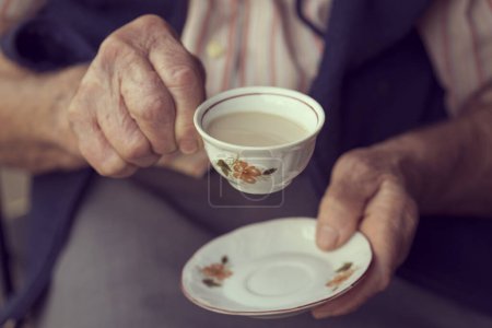 Téléchargez les photos : Détail des mains d'une femme âgée tenant une tasse rustique remplie de café à la crème - en image libre de droit