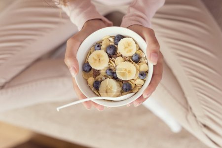Photo for Detail of a young woman sitting on a living room couch, holding a bowl of cereal and having breakfast. Selective focus - Royalty Free Image