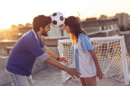 Photo for Young couple in love having fun on a building rooftop after playing football, dancing and balancing with the ball between their heads - Royalty Free Image