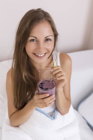 Photo for Beautiful girl sitting on a bed after waking up in the morning, smiling and holding a glass of raspberry and blueberry mix smoothie and smiling - Royalty Free Image