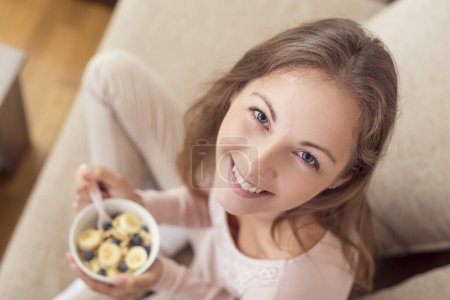 Photo for Top view of a young woman sitting on a living room couch, holding a bowl of cereal and having breakfast - Royalty Free Image