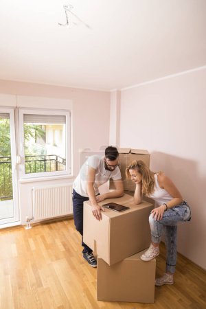 Photo for Young couple in love moving in a new apartment, standing next to cardboard boxes, holding tablet computer and surfing the web while planning to redecorate their new home - Royalty Free Image