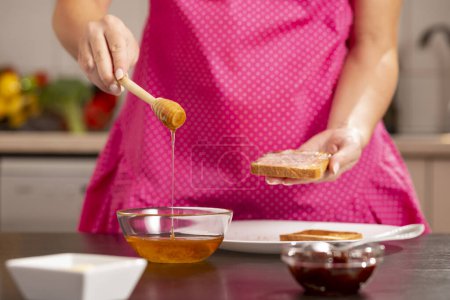 Téléchargez les photos : Détail des mains féminines tenant une tranche de pain grillé et une cuillère en bois, étalant du miel sur le pain ; femme faisant un sandwich au beurre et au miel pour le petit déjeuner - en image libre de droit
