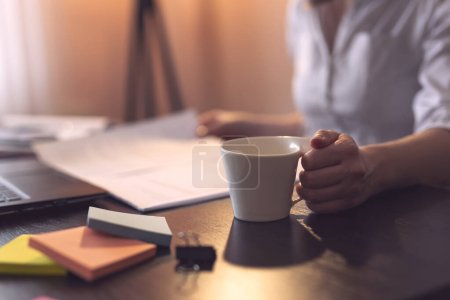 Téléchargez les photos : Femme d'affaires assise à son bureau dans un bureau à domicile, examinant un contrat et buvant du café. Focus sélectif sur le pouce et la tasse - en image libre de droit