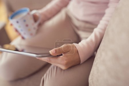 Foto de Detalle de una hermosa joven sentada en un sofá de la sala de estar, sosteniendo una tableta, una taza de café y navegando por la red - Imagen libre de derechos