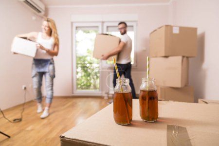 Photo for Young couple in love carrying cardboard boxes and moving in their new apartment. Two ice tea bottles with drinking straws placed on one of the cardboard boxes in focus - Royalty Free Image