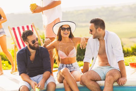 Foto de Grupo de amigos en una fiesta de verano junto a la piscina, sentados al borde de una piscina, tomando cócteles y divirtiéndose - Imagen libre de derechos