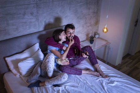 Téléchargez les photos : Beau jeune couple amoureux, assis au lit côte à côte, étreignant, mangeant du pop-corn et s'amusant à regarder une émission de télévision la nuit - en image libre de droit