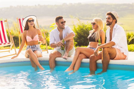 Foto de Grupo de amigos en una fiesta de verano junto a la piscina, sentados al borde de una piscina con piernas en el agua, bebiendo cerveza y divirtiéndose - Imagen libre de derechos