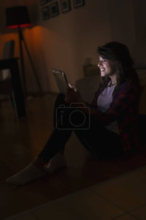 Téléchargez les photos : Belle jeune femme à la maison, assise dans le noir d'un salon, surfant sur le net sur une tablette, profitant de son temps libre - en image libre de droit
