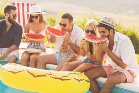Photo for Group of friends at a poolside summer party, sitting at the edge of a swimming pool, eating cold watermelon slices and having fun - Royalty Free Image