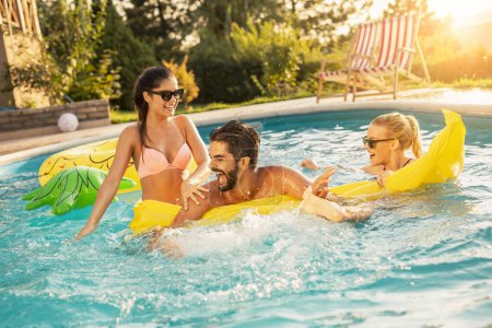 Téléchargez les photos : Groupe d'amis à une fête d'été au bord de la piscine, s'amuser dans la piscine, éclabousser de l'eau et se battre sur un matelas flottant - en image libre de droit