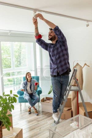Photo for Couple in love moving in together, man standing on the ladder and changing the light bulb while woman is drinking coffee and relaxing - Royalty Free Image