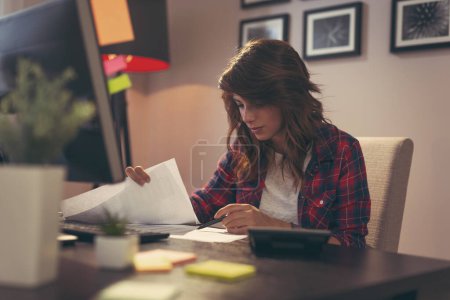 Téléchargez les photos : Jeune femme examinant les documents, travaillant tard dans un bureau à domicile - en image libre de droit