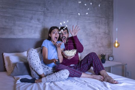 Téléchargez les photos : Beau jeune couple amoureux, assis au lit côte à côte, mangeant du pop-corn et s'amusant à regarder un film d'horreur la nuit - en image libre de droit