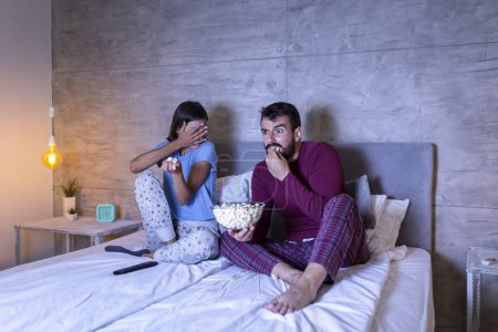 Foto de Hermosa pareja joven enamorada, sentada en la cama lado a lado, comiendo palomitas de maíz y divirtiéndose viendo una película de terror por la noche - Imagen libre de derechos