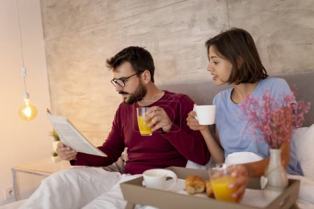 Foto de Hermosa pareja joven acostada en la cama por la mañana, desayunando y leyendo periódicos - Imagen libre de derechos