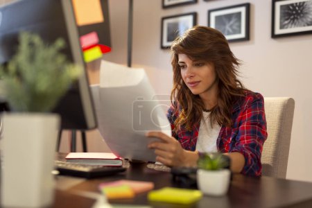 Téléchargez les photos : Jeune femme assise à un bureau à son bureau à domicile, tenant et examinant certains documents, faisant des heures supplémentaires - en image libre de droit