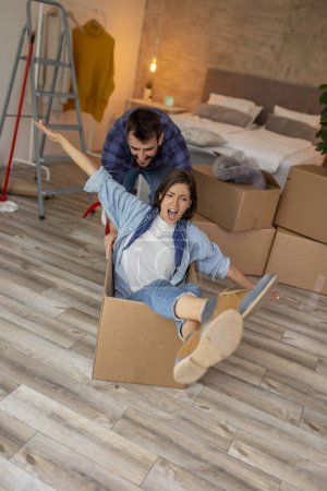 Photo for Beautiful young couple in love moving in together, having fun while unpacking cardboard boxes with their belongings, guy pushing girl around in a cardboard box - Royalty Free Image