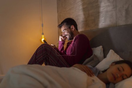 Photo for Man using smart phone while his girlfriend is asleep in bed at night; insomnia and sleeping disorders concept - Royalty Free Image