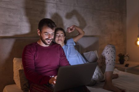 Photo for Beautiful couple lying in bed at night, using a laptop computer and relaxing at home after a busy day - Royalty Free Image