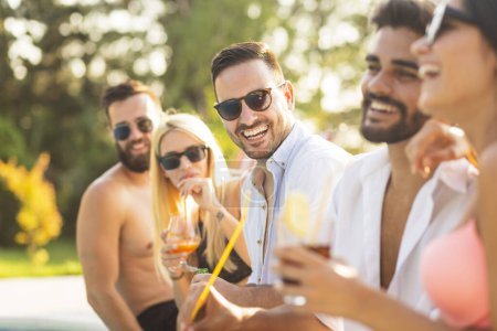 Foto de Grupo de amigos en una fiesta de verano junto a la piscina, sentados al borde de una piscina, bebiendo cócteles y cerveza y divirtiéndose - Imagen libre de derechos