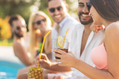 Foto de Grupo de amigos divirtiéndose en una fiesta de verano junto a la piscina, sentados al borde de una piscina con piernas en el agua, bebiendo cerveza y cócteles - Imagen libre de derechos