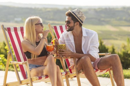 Téléchargez les photos : Couple amoureux à une fête d'été au bord de la piscine, assis sur des chaises longues au bord de la piscine, boire des cocktails et s'amuser - en image libre de droit