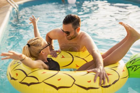 Foto de Pareja enamorada en una fiesta de verano junto a la piscina, tomando el sol y divirtiéndose; chica flotando en la piscina en un colchón de aire en forma de piña - Imagen libre de derechos