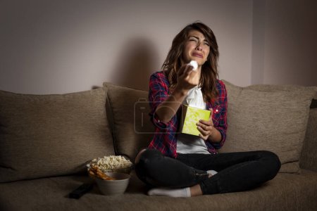 Photo for Beautiful young woman sitting in the dark on a living room couch, watching a soap opera and crying - Royalty Free Image