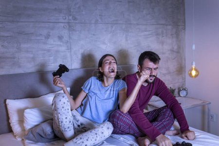 Photo for Beautiful young couple wearing pajamas, having fun at home, playing video games in bed at night, woman excited after winning the game, man disappointed after losing - Royalty Free Image