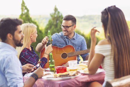 Photo for Group of friends having a barbecue party outdoors, drinking beer, grilling meat, playing the guitar, singing and having fun - Royalty Free Image