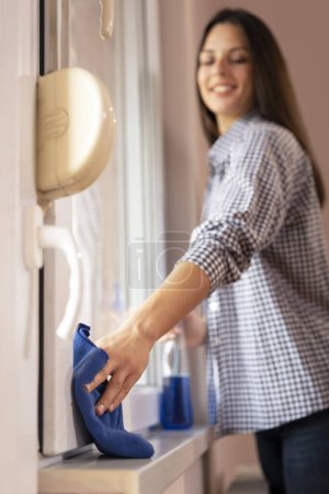 Téléchargez les photos : Belle jeune femme nettoyant les fenêtres avec un spray nettoyant et un chiffon ; aide à l'entretien ménager essuyant les fenêtres. Focus sélectif sur la main tenant le tissu - en image libre de droit
